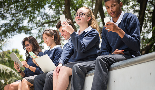 Group of five students enjoying their phones and laptops outdoors.