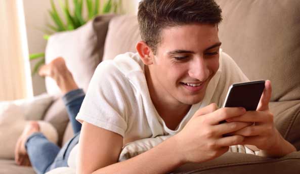 A young man laying comfortably on the sofa, happily smiling whilst on his phone